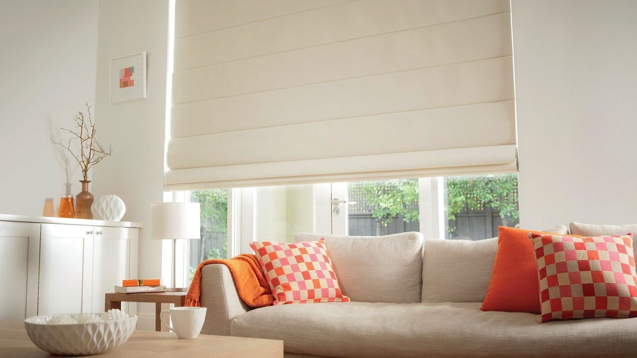 A living area with a cream couch, orange cushions and white Roman blinds.