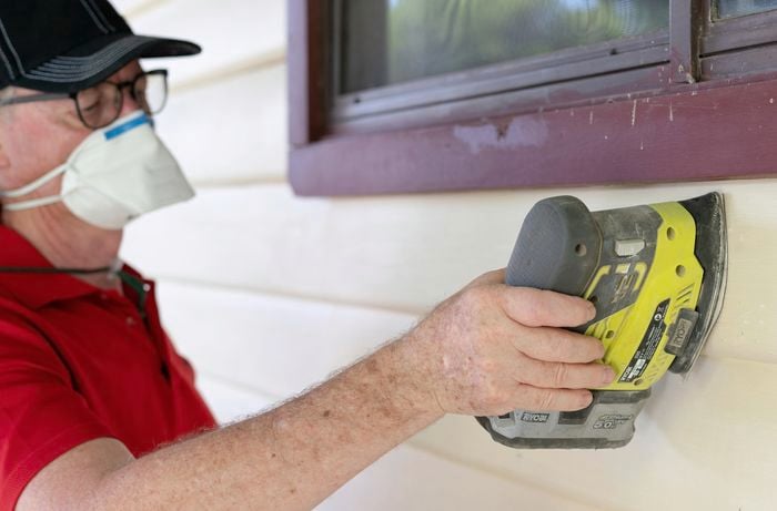 A person wearing a mask sanding weatherboards with a cordless sander