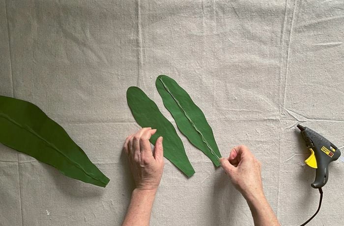 Hands positioning wire on bead of glue on a green leaf cut-out, with a hot-glue gun nearby.