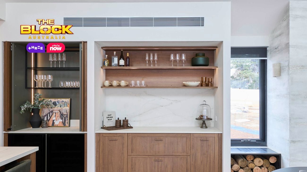 Modern kitchen bar area from The Block, featuring wooden cabinetry, open shelves with glassware and decor, a built-in wine fridge, glass cabinet, and a firewood storage nook beside a large window.