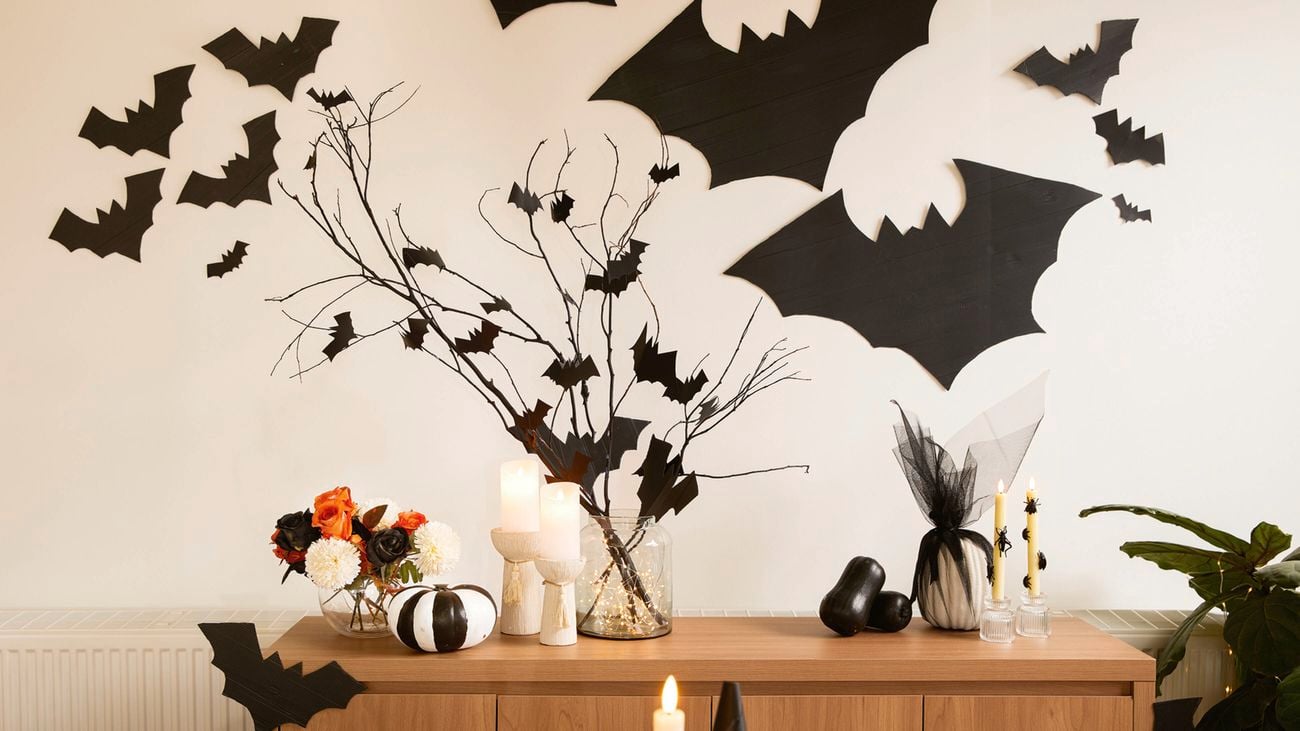 A wooden cabinet with Halloween decorations, including black bat cut-outs, white and black pumpkins, candles and a vase with branches. More bat cut-outs are on the wall above.