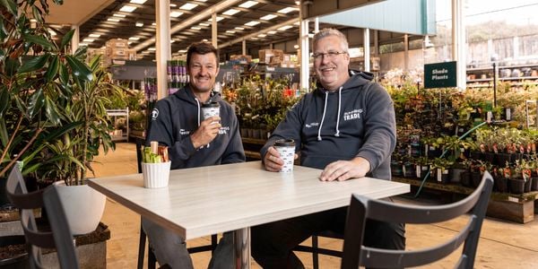 Two males sitting at a table holding cups of coffee with plants around them.