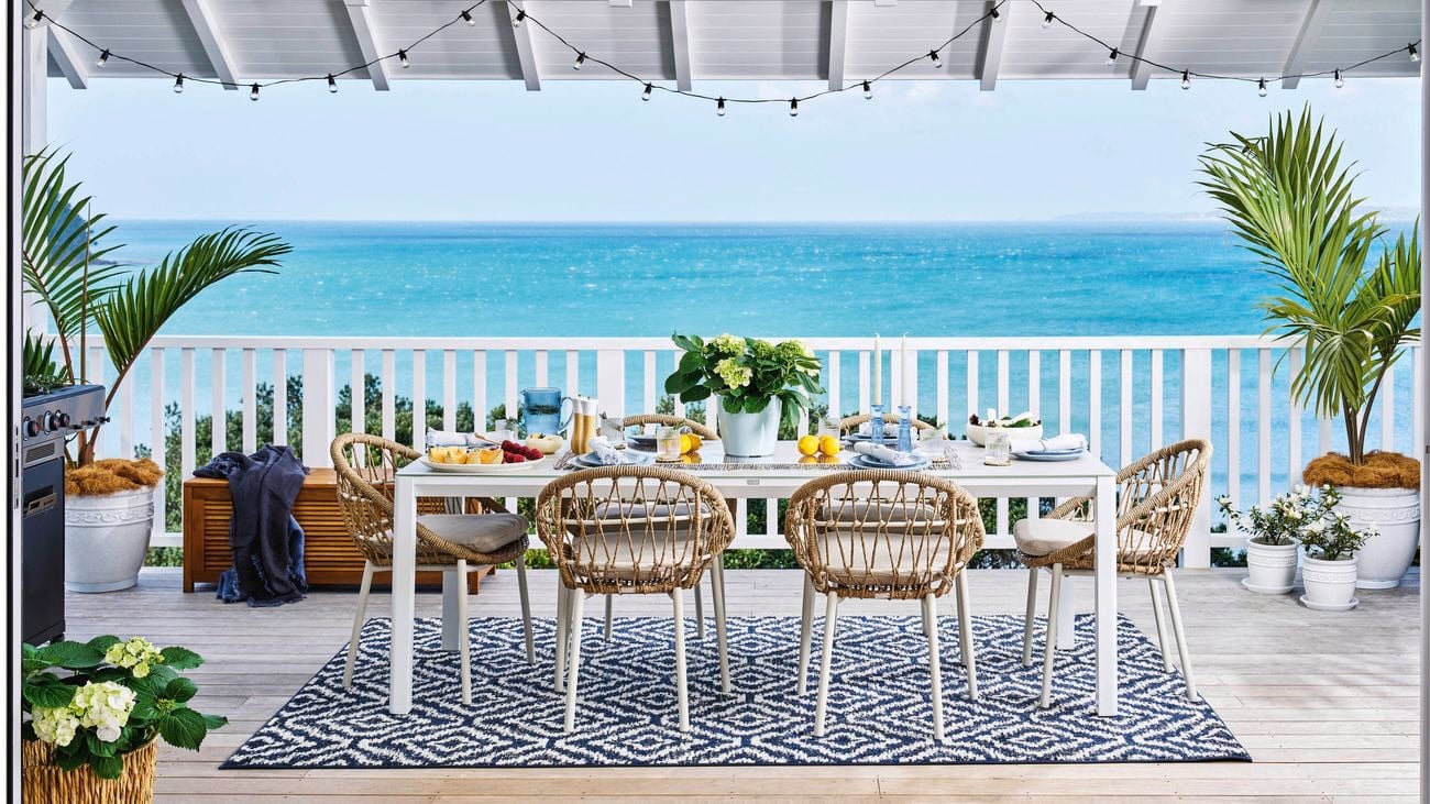   Outdoor dining setup on a wooden deck overlooking the ocean, featuring a white rectangular table with wicker chairs, a patterned black-and-white rug, potted plants, and string lights hanging from a white pergola roof.