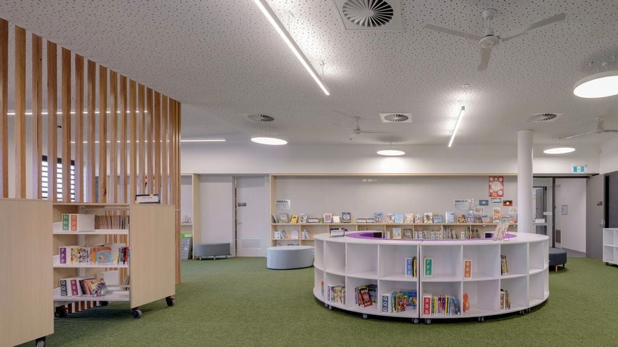 Interior of a modern library with green carpet, circular bookshelf, wooden partitions, and ceiling fans under white perforated ceiling tiles