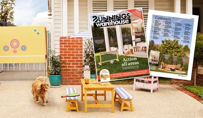 Dog carrying a toy in a family-friendly front yard play space with a tennis net, outdoor table and benches, while Bunnings Warehouse magazine cover and contents pages are displayed in the foreground.