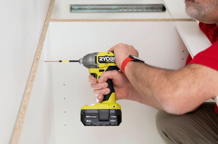 A person is using a power drill to attach a base cabinet panel to the wall.