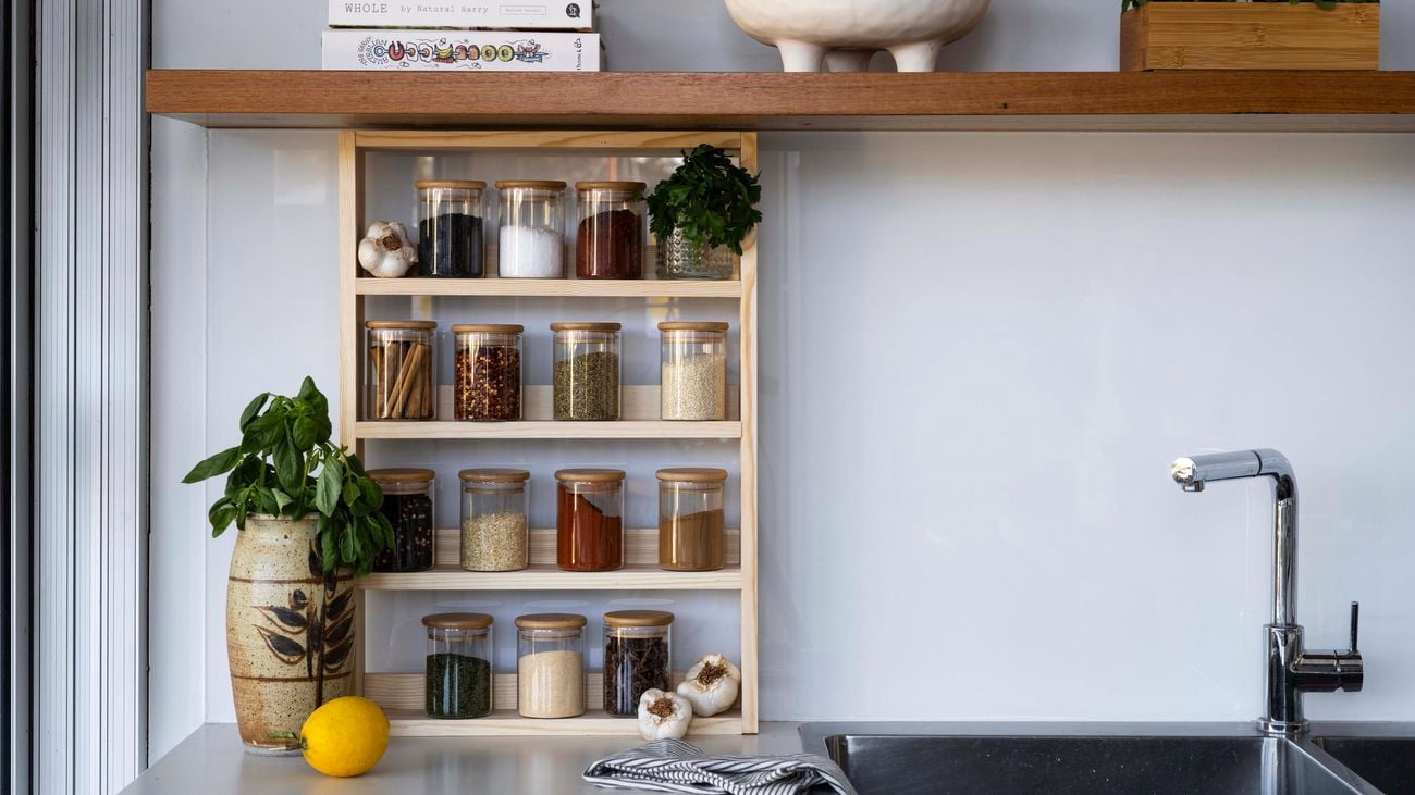 A wooden spice rack with jars of spices in it on a kitchen bench next to a sink