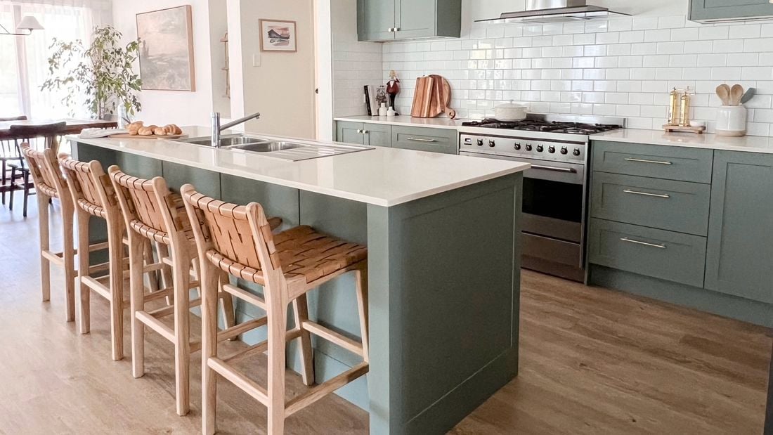 A modern kitchen with green cupboards, marble benchtop, with bar stools at the bench and timber flooring. 