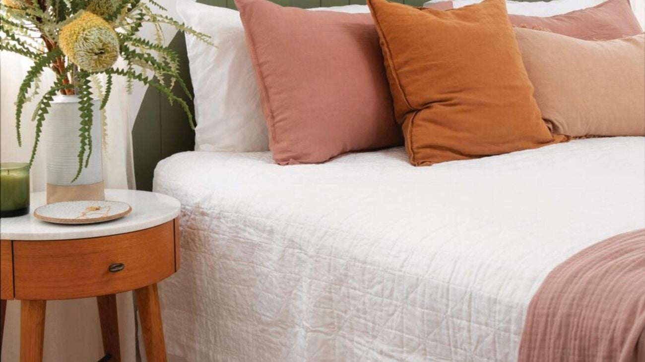 Bed with white covers, green arched headboard, pink and orange pillow and a side table with flower vase on top