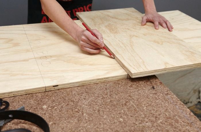A person drawing a line on a plywood sheet using another sheet as a straight edge