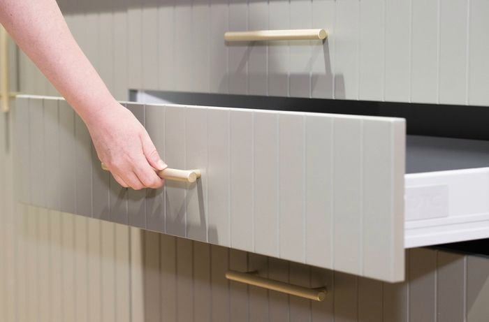 A person with their hand on a kitchen drawer gold handle with the drawer open in a kitchen.