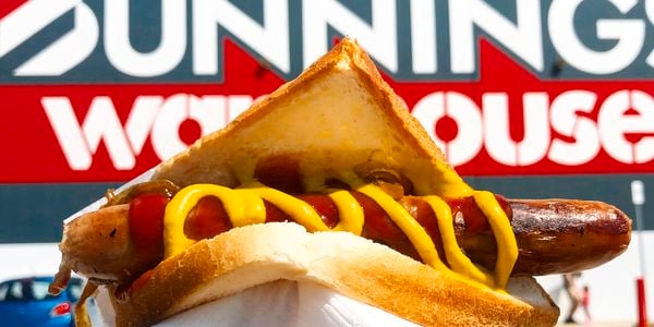 Hand holding a Bunnings sausage in bread with tomato sauce and mustard, in front of a large Bunnings Warehouse storefront.