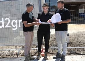 Three workers in work boots and dark shirts stand on a footpath next to a temporary construction fence, holding a large paper plan together. Muddy ground and a partially built brick house are behind the fence.]