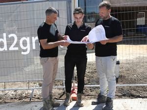 Three workers in work boots and dark shirts stand on a footpath next to a temporary construction fence, holding a large paper plan together. Muddy ground and a partially built brick house are behind the fence.]