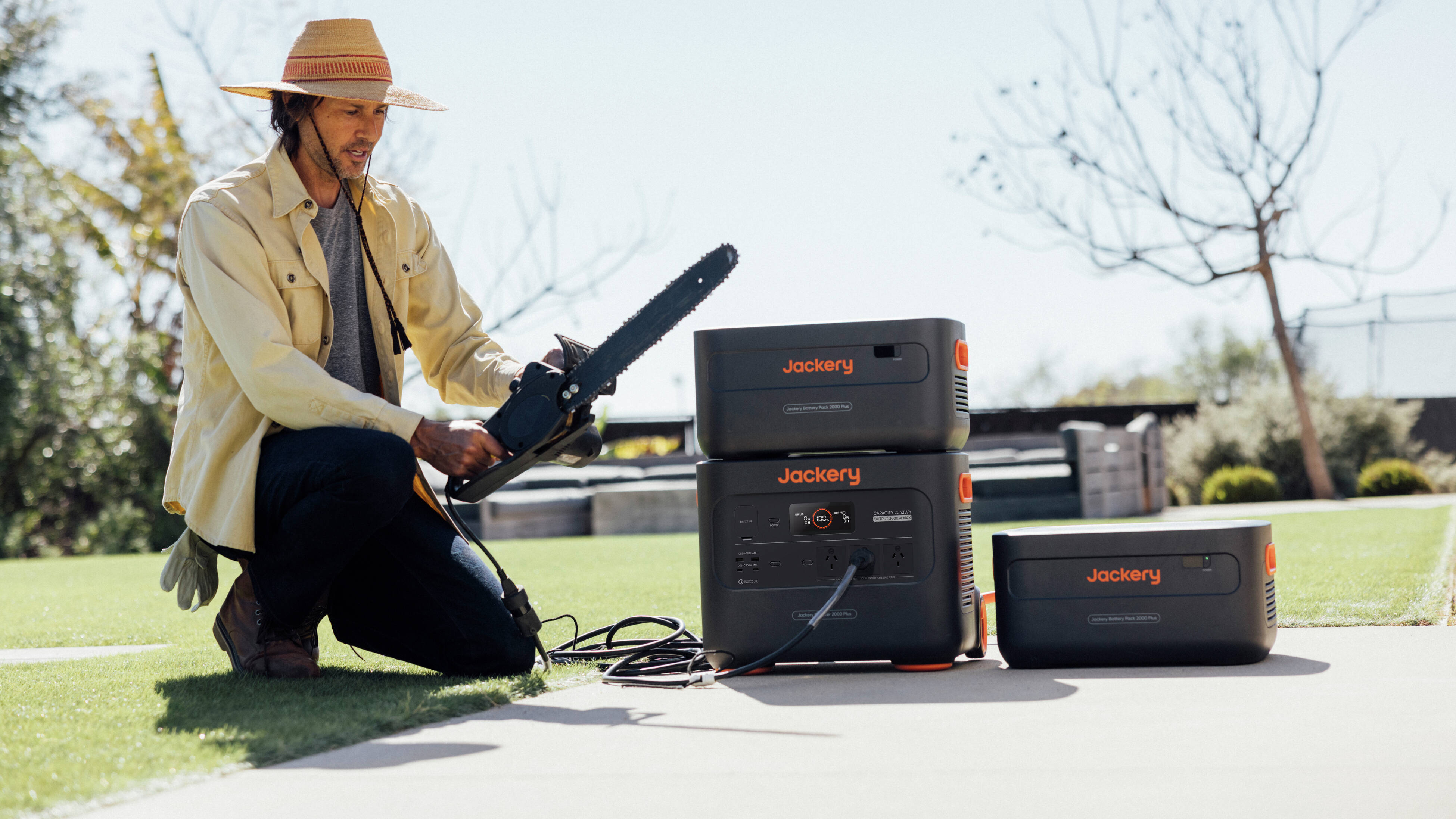 : A man, wearing a wide-brimmed hat, white long sleeved shirt and holding a chainsaw in his hand which is connected to a Jackery portable power station.