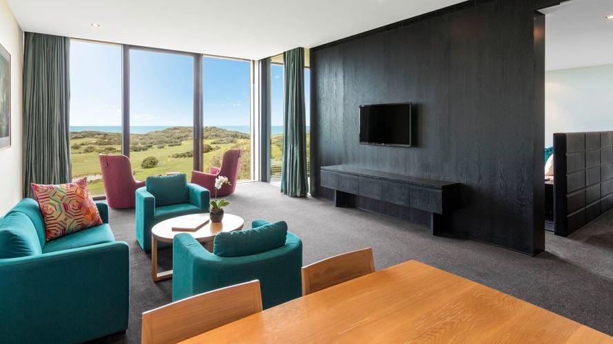 Modern living room with teal armchairs, pink chairs by large windows overlooking greenery and the ocean, a wooden coffee table, wall-mounted TV, and a glimpse of an adjacent room through an open doorway