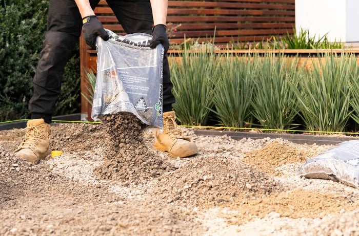 A person wearing gloves and work boots is spreading road base from a bag across the cleared area