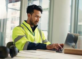 A man in a high-vis vest works on a laptop at a desk with construction plans and a hard hat