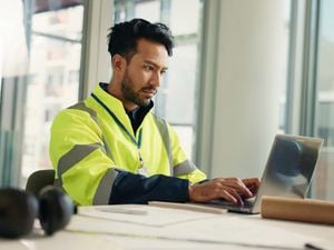 A man in a high-vis vest works on a laptop at a desk with construction plans and a hard hat