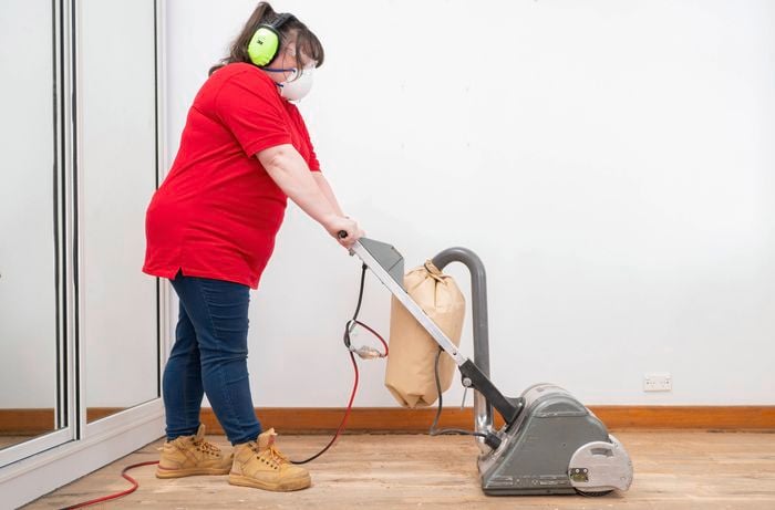 A person wearing safety gear is sanding a timber floor with a drum sander, maintaining a steady pace and overlapping passes for an even surface. 