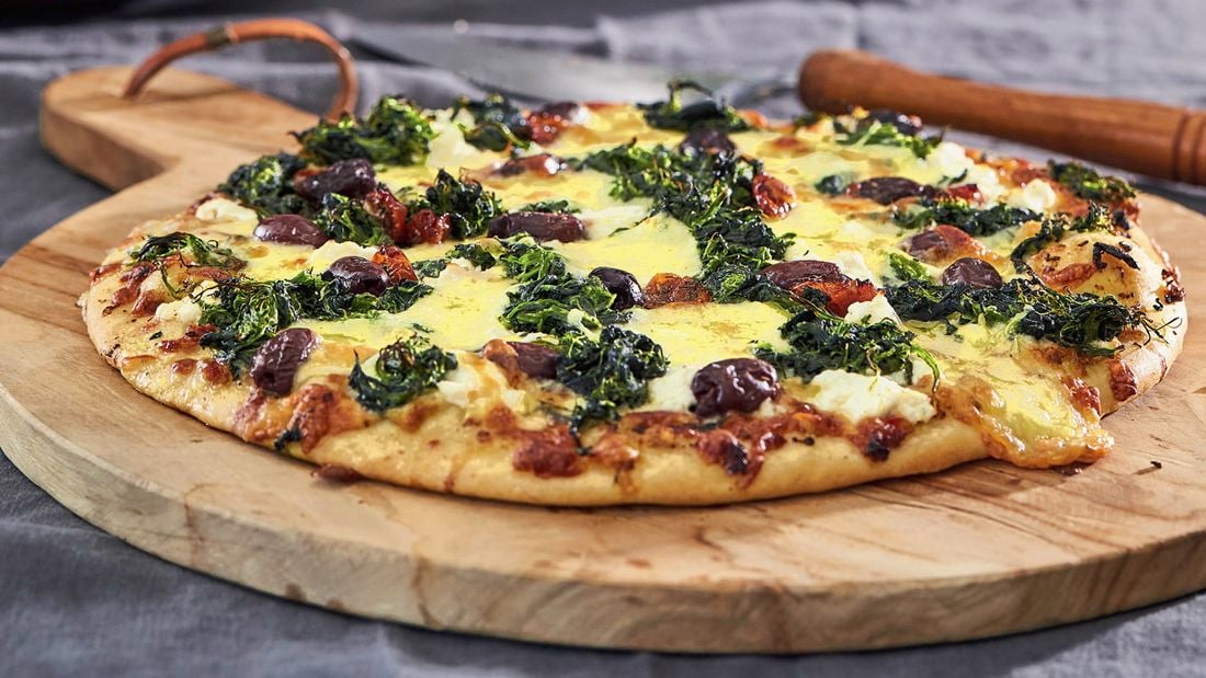 A cooked spinach and feta cheese pizza with olives served on a round wooden board with handle
