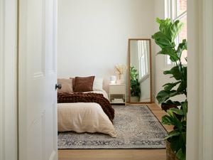 A calm bedroom viewed through a doorway, featuring a neatly made bed with neutral bedding, a patterned rug, a bedside table, a full-length mirror, and a large indoor plant by a window.