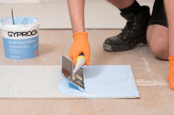 A person wearing orange gloves is spreading blue Gyprock acrylic stud adhesive on the back of sheet using a notched trowel.