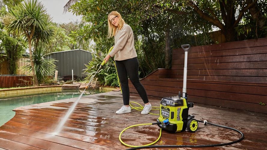 A woman wearing safety glass and using a pressure washer to clean deck of a house.