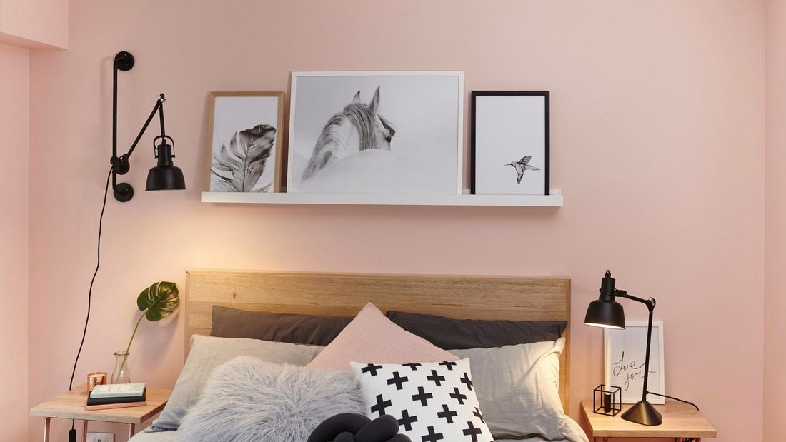 A floating white shelf with framed pictures above a bedhead against a pink wall