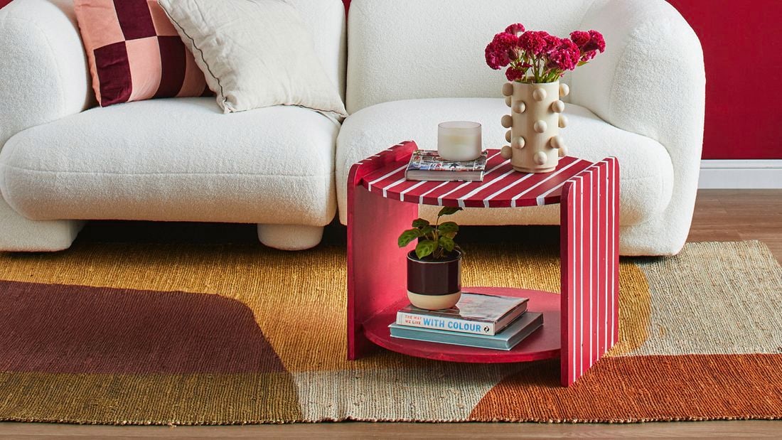 A red and white striped two tier coffee table with a vase, candle and book on top next to a white lounge