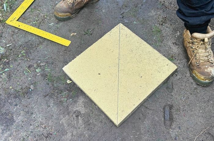 A single paver on top of dirt with part of a large yellow framing square and a person’s boot in image