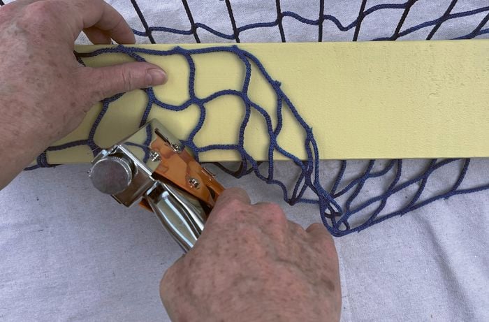 : Hands using a staple gun to attach a blue net to a yellow piece of timber