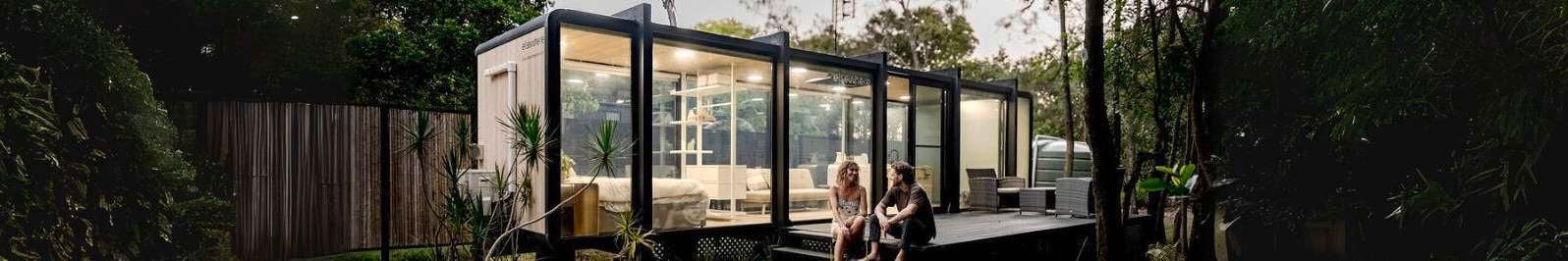 Two people sitting on decking outside a room with lights on.