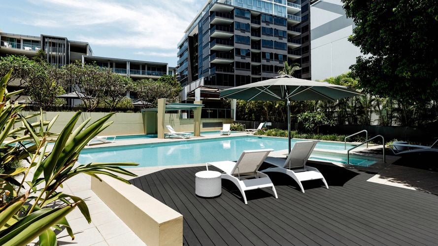 Modern outdoor swimming pool area with two white lounge chairs and a small round table under a large umbrella, surrounded by greenery and tall residential buildings