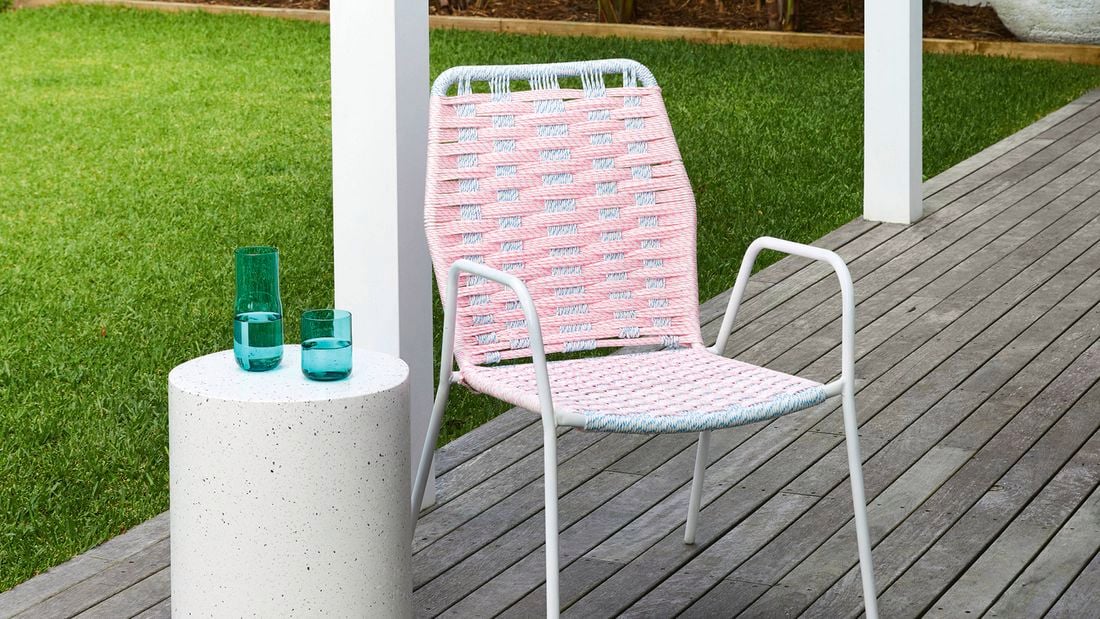 A white patio chair repaired with vibrant rope using D.I.Y. outdoor chair weaving techniques, sitting on a timber deck with a green grassy background and a white side table holding a glass of water.