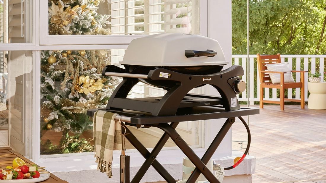 A portable single‑burner gas barbecue on a foldable trolley with a gas bottle sits on a timber deck verandah beside a window; a decorated Christmas tree is visible inside, with outdoor furniture and trees in the background.