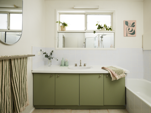 Bright bathroom with a green vanity cabinet and white benchtop, featuring a central sink beneath a wide mirror, a bathtub to the side, white tiled walls, striped towel on a rack, and potted plants along the windowsill for natural light and greenery.