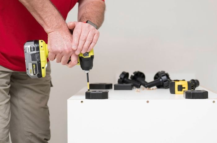 A person is using a power drill to attach adjustable feet to the bottom of a base cabinet. 