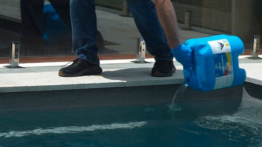 Person wearing blue gloves pouring liquid from a blue container into a swimming pool.