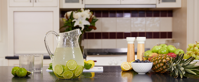 Glass jug half filled with water with slices of lime on a bench with glasses and fruit