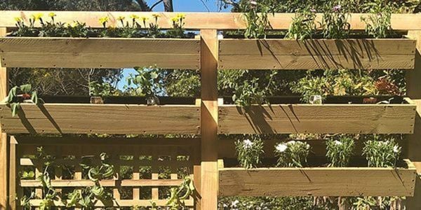 A vertical wooden garden structure with multiple planter boxes holding green plants and flowers, set outdoors among lush greenery.