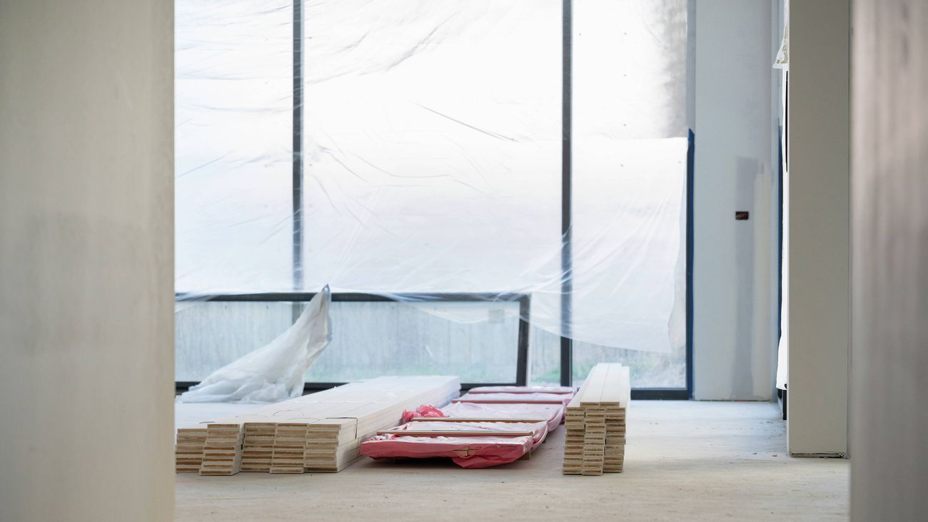 Interior room under construction with stacked timber flooring boards on a concrete floor, large windows covered with protective plastic sheeting, and unfinished wall surfaces awaiting final fit-out.