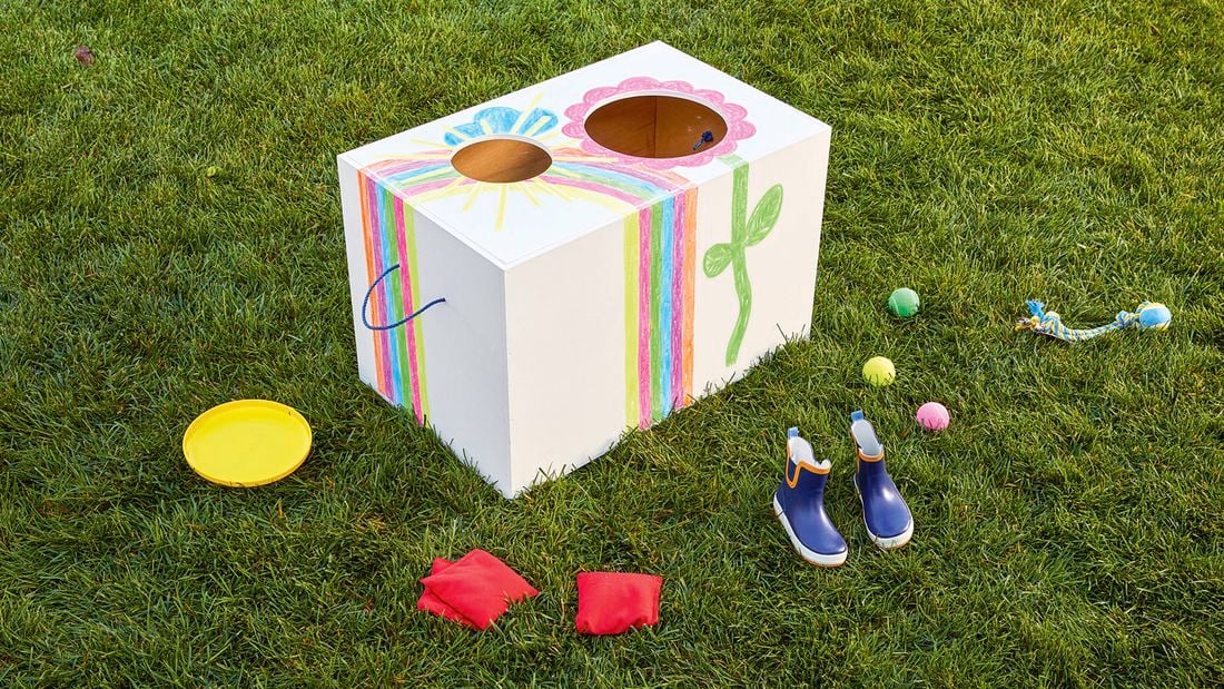 A white wooden toy box decorated with colourful drawings sitting on grass, surrounded by toys and gumboots