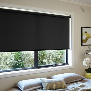 A black roller blind installed in a window in a light themed bedroom