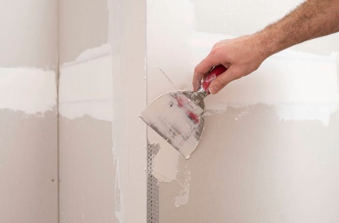 A hand is applying a base coat of plaster over the corner bead with a trowel. 