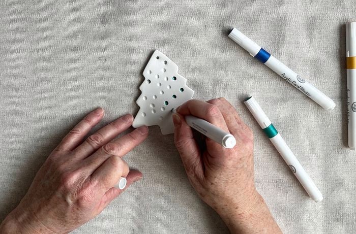 A pair of hands holding a marker pen and a pen lid colouring a white clay Christmas tree on a tablecloth; metallic marker pens sit to the right.