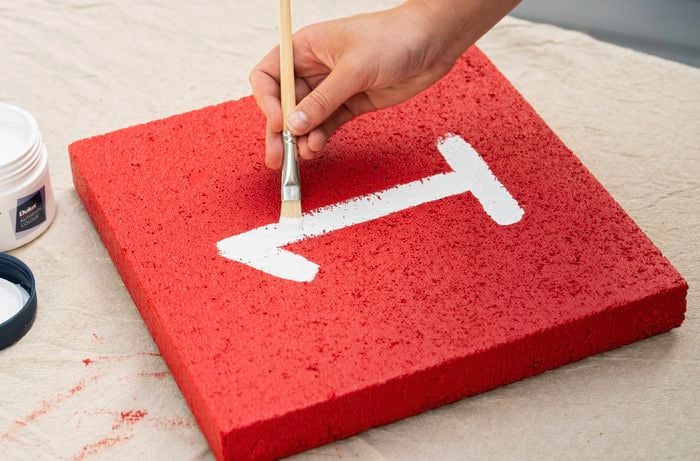 A person is using a paint marker to draw a number on a red painted paver, part of a reusable hopscotch path.