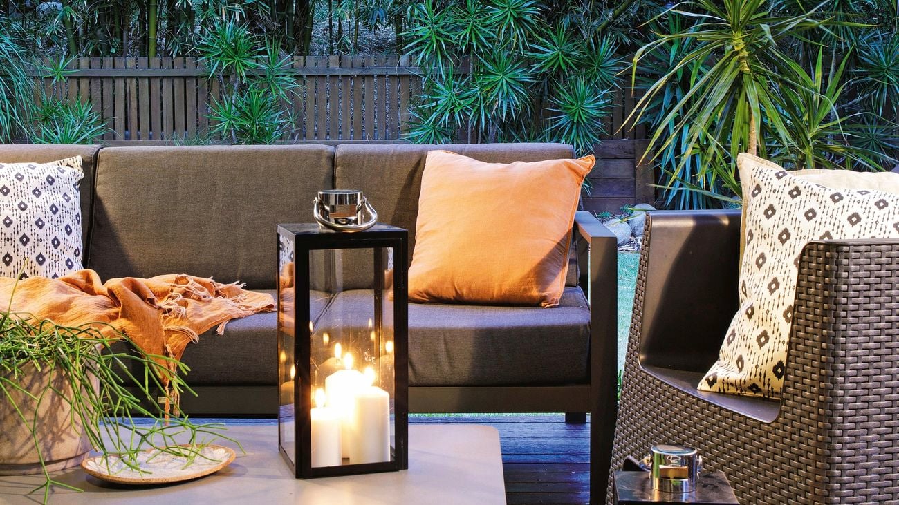 Outdoor lounge and armchair seated around outdoor table with two lamps in foreground on top of synthetic grass.