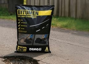 A 20kg bag of bitumen placed on a road next to a pothole, with grass and a wooden fence in the background