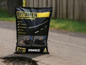 A 20kg bag of bitumen placed on a road next to a pothole, with grass and a wooden fence in the background